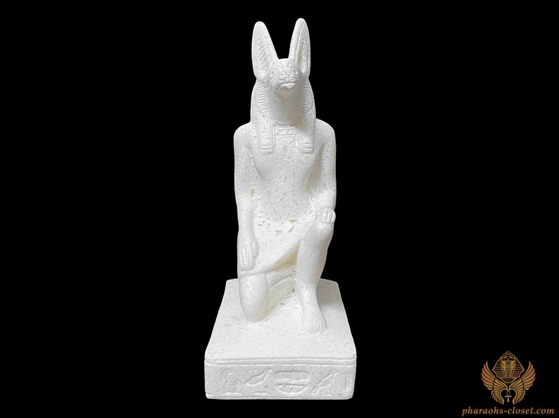 Puede incluir: Estatua de cer&aacute;mica blanca de un dios egipcio con cabeza de chacal, arrodillado sobre una base con jerogl&iacute;ficos. La estatua es detallada y tiene una superficie texturizada.