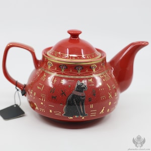 Op de afbeelding: Rode keramische theepot met een zwarte kat en gouden Egyptische hiërogliefen. De theepot heeft een deksel en een tuit.