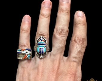 May include: Two silver rings with colourful stones. The ring on the index finger features a blue and turquoise scarab beetle design. The ring on the middle finger features a blue and orange eye of Horus design.
