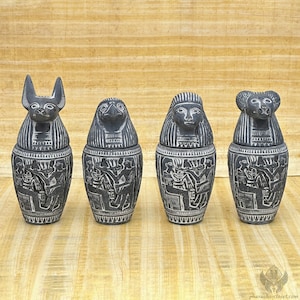 Op de afbeelding: Vier zwarte en witte keramische canopische potten met Egyptische hiërogliefen en dierlijke deksels. De potten zijn in een rij op een lichtbruin oppervlak gerangschikt.