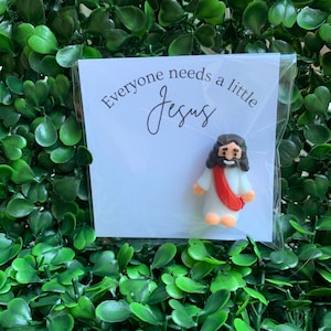 May include: A small figurine of a person with brown hair and a beard, wearing a white robe with a red sash, attached to a white card. The card features the black printed text "Everyone needs a little Jesus" in a script and sans-serif font.