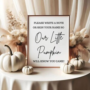 May include: A framed sign with the text "PLEASE WRITE A NOTE OR SIGN YOUR NAME SO Our Little Pumpkin WILL KNOW YOU CAME!" is surrounded by white pumpkins and floral arrangements. The sign is in a gold frame.