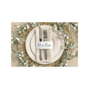 May include: A formal place setting with a white plate, a folded beige napkin, and silverware. A white card with the text "He is Risen" is placed on the napkin. The setting is decorated with a gold-colored placemat and greenery.