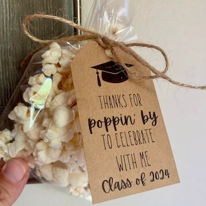 Op de afbeelding: Een bruine papieren cadeauzak met een afstudeerhoedontwerp en de tekst "Bedankt voor het langskomen om met me te vieren Klas van 2024". De zak is gevuld met popcorn en vastgebonden met touw.