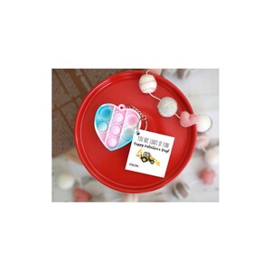 May include: A heart-shaped pop-it toy in shades of blue, pink, and white, attached to a keychain. The toy sits on a red plate, next to a small card that reads "YOU ARE LOADS OF FUN! Happy Valentine's Day!" with a yellow tractor graphic.