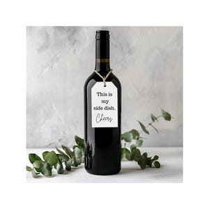 May include: A black wine bottle with a white tag that reads "This is my side dish. Cheers." The tag is tied with twine. Green leaves are in the background.