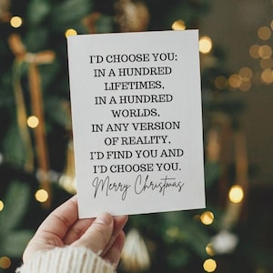 Könnte beinhalten: Eine weiße Karte mit schwarzem Text, gehalten in einer Hand, vor einem verschwommenen Hintergrund aus Weihnachtslichtern. Auf der Karte steht: "I'D CHOOSE YOU; IN A HUNDRED LIFETIMES, IN A HUNDRED WORLDS, IN ANY VERSION OF REALITY, I'D FIND YOU AND I'D CHOOSE YOU. Merry Christmas."
