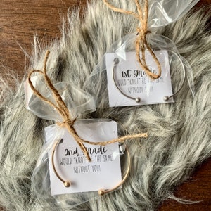 Puede incluir: Dos pulseras de puño abiertas, una plateada y otra dorada, empaquetadas en celofán transparente con lazos de cuerda. Cada pulsera tiene una pequeña bola en cada extremo. Las tarjetas dicen "1st grade would 'knot' be the same without you!" y "2nd grade would 'knot' be the same without you!"
