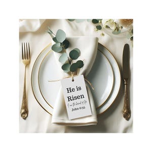 May include: Elegant table setting with a white napkin, plate, and gold-rimmed accents. A tag with the text "He is Risen" and John 6:35 is tied to the napkin. Eucalyptus sprigs add a natural touch. A gold fork and knife complete the arrangement.