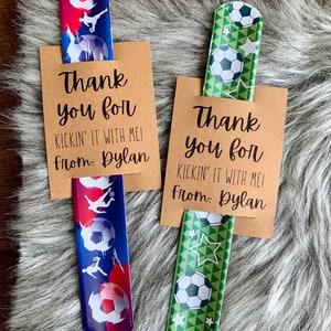 May include: Two brown cardstock thank you tags with the text "Thank you for kickin' it with me! From: Dylan" and a soccer-themed stretchy bracelet with a blue and white design and a green and white design.