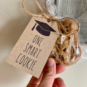 May include: A brown paper gift tag with a black graduation cap and the text "One Smart Cookie". A cookie is tied to the tag with twine.