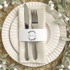 May include: A place setting with a white plate, a beige napkin, and silverware. A white paper band with the text "Jake & Cari" and a leaf design encircles the napkin, knife, and fork. Decorative greenery and a gold placemat surround the plate.