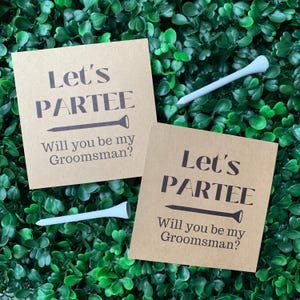 Pode incluir: Dois cartões marrons com o texto "Let's Partee" e "Will you be my Groomsman?" impresso neles. Cada cartão tem um gráfico de tee de golfe ao lado do texto.