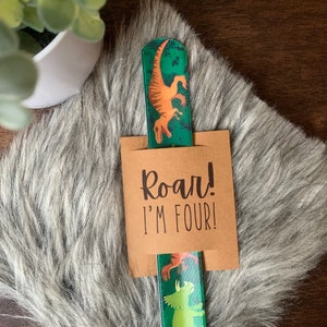 May include: A green and brown dinosaur-themed bookmark with the text "Roar! I'm Four!" printed on a brown card. The bookmark features illustrations of dinosaurs in orange and green. The background is a faux fur surface.