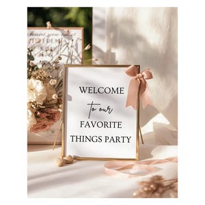 Może przedstawiać: Prostokątny znak z napisem "WELCOME to our FAVORITE THINGS PARTY" w czarnym skrypcie, w złotej ramie. Różowa kokarda zdobi ramę. Tło zawiera kwiaty i kolejny oprawiony znak.
