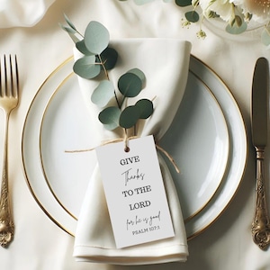 Puede incluir: Una elegante mesa con una servilleta blanca, un plato y un plato con borde dorado. Una etiqueta con el texto "GIVE Thanks TO THE LORD for he is good PSALM 107:1" está atada a la servilleta con hilo. Ramas de eucalipto añaden un toque natural. Un tenedor y un cuchillo dorados completan el arreglo.
