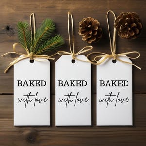 Pode incluir: Três etiquetas de presente brancas com as palavras "BAKED with love" impressas em preto. Cada etiqueta é amarrada com uma fita dourada e adornada com um raminho de sempre-viva ou pinhas, sobre um fundo de madeira.