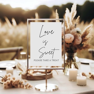 Peut inclure: Un panneau doré avec l'inscription "Love is Sweet" en écriture élégante, avec "PLEASE TAKE A TREAT" en dessous. Le panneau est sur un support doré, posé sur une table avec des bougies et des compositions florales, créant une ambiance romantique.