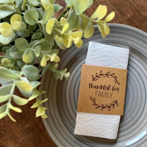 May include: A brown paper napkin holder with a white napkin inside. The holder has a brown background with a wreath of leaves and the text "thankful for FAMILY" in black.