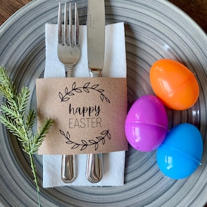 May include: A brown paper place card with the text "happy easter" in black lettering surrounded by a wreath of leaves. The place card is wrapped around a fork and knife on a white napkin. Three plastic Easter eggs are on a gray and white striped plate. The eggs are orange, purple, and blue.