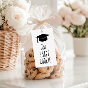 May include: A clear plastic bag filled with chocolate chip cookies, tied at the top with a light beige ribbon. A white rectangular tag hangs from the ribbon, featuring a black graduation cap icon and the text ONE SMART COOKIE in black.