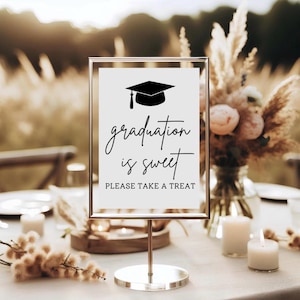 May include: A graduation sign with a black mortarboard illustration and the words "graduation is sweet, please take a treat" in elegant script. The sign is in a gold frame, set on a table with candles and floral arrangements.