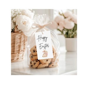 May include: A clear cellophane bag tied with a beige ribbon, filled with chocolate chip cookies. A white tag with the words "Happy Easter" and a watercolor bunny illustration is attached to the bag. A wicker basket and white flowers are in the background.