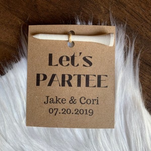May include: A brown card with the text "Let's Partee" in black, followed by "Jake & Cori" and "07.20.2019". A white golf tee is attached to the top of the card.