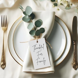 Puede incluir: Una tarjeta de lugar blanca con caligrafía negra que dice "Thankful For Family" atada con un cordel a una servilleta blanca con una rama de eucalipto.