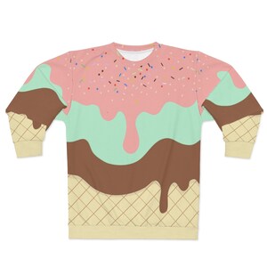 Op de afbeelding: Een sweatshirt met lange mouwen met een kleurrijk ijshoorntjesontwerp. Het sweatshirt heeft roze, groene en bruine ijs met sprankelingen erop, alles smelt over een wafelhoorn.