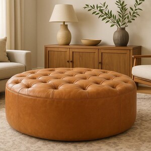 May include: A large, round, tufted ottoman in a warm, caramel-colored leather. The ottoman is the focal point of a living room setting, surrounded by a sofa, armchair, and wooden cabinet. The room has a neutral color palette.
