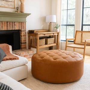 May include: A living room scene featuring a large, round, tufted ottoman in a warm brown leather. The room includes a white sofa, a wooden console table with woven baskets, and a brick fireplace. A woven chair sits near a window.