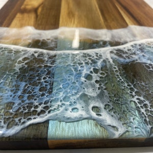 May include: A wooden cutting board with a wave design made of white and blue resin. The wave has a foamy texture and is set into the wood.
