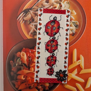 May include: A handmade bookmark featuring a cross-stitched design of four red ladybugs with black spots on a white background. The bookmark has a red polka dot ribbon at the top and a black flower at the bottom. The bookmark is displayed next to pasta dishes.