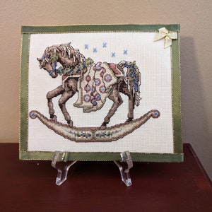 May include: A white cross-stitch of a rocking horse with a green and brown blanket. The horse is on a white background with a green border and a yellow ribbon.