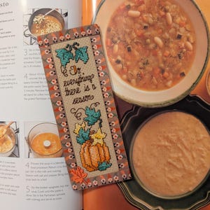 May include: A handmade bookmark with a cross-stitched pumpkin design and the text "Do everything there is a season." The bookmark has a tan background with a decorative border. The image also shows bowls of soup and a cookbook.