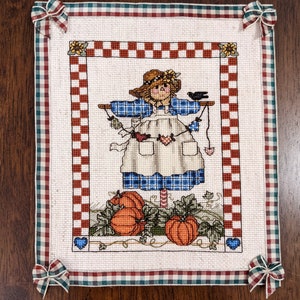 May include: A cross-stitch embroidery of a scarecrow with a blue and white plaid shirt, a white apron, and a straw hat. The scarecrow is holding a string of hearts and is surrounded by pumpkins and leaves.