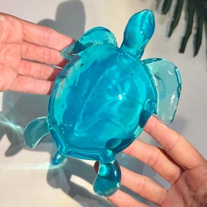 Könnte beinhalten: Eine türkisblaue Glasfigur einer Meeresschildkröte. Die Schildkröte sitzt auf einer weißen Oberfläche.
