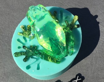 May include: A green glass frog figurine in a blue silicone mold. The mold has a textured surface that resembles the frog's skin. The frog is translucent and has a shiny finish. The mold is round and has a flat bottom. The text "@frökenhögnert" is visible in the lower right corner of the image.