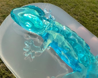 May include: A clear blue resin gecko figurine in a silicone mold. The gecko is detailed with textured skin and has a translucent appearance.
