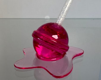 May include: A pink lollipop with a white stick, melting into a puddle on a reflective surface.