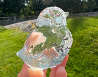 May include: A clear glass bird figurine with a rounded shape and intricate details. The figurine is held in a hand against a green grassy background.