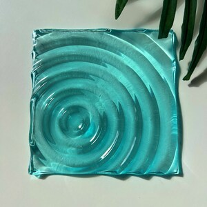 Puede incluir: Un posavasos cuadrado de vidrio azul claro con un diseño ondulado que parece agua. El posavasos tiene una forma ligeramente irregular.