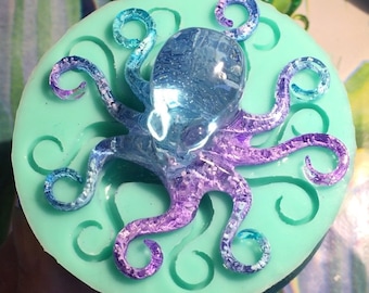 May include: A teal blue silicone mold shaped like an octopus with eight tentacles. The mold is filled with a clear, iridescent purple and blue resin octopus.