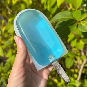 Może przedstawiać: Niebieski element artystyczny z żywicy w kształcie loda na patyku ze srebrnym brokatowym patyczkiem. Żywica ma efekt gradientu, przechodząc od ciemniejszego turkusu do jaśniejszego błękitu. Lodzik jest w białej formie.