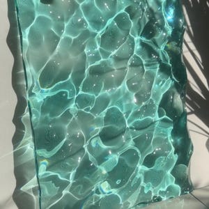 May include: A rectangular piece of translucent teal glass with a textured, rippled surface that mimics the appearance of moving water. The glass features light-refracting patterns and deep blue-green hues, creating a dynamic visual effect.