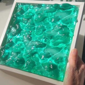 May include: A square, white-framed artwork featuring a textured, translucent, teal-colored resin design. The resin appears to have a rippled surface, creating a water-like effect. The artwork is held by a hand.