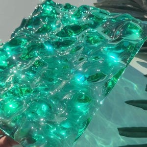 May include: A rectangular sheet of translucent emerald green glass featuring a textured, rippled surface that mimics the appearance of moving water. The glass catches the light, creating bright highlights and deep green shadows across its surface.