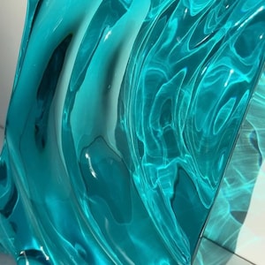 May include: Abstract turquoise glass sculpture with a rippled, wave-like design. The translucent material reflects light, creating highlights and shadows. The artwork has a modern aesthetic, suitable for home decor.