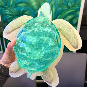 Puede incluir: Una figurita de tortuga marina de vidrio verde azulado con una concha blanca texturizada. La tortuga está sentada sobre una superficie blanca.